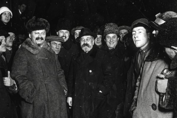 Fotografia histórica em preto e branco mostrando um grupo de homens em um ambiente noturno com neve caindo. Eles vestem casacos pesados e chapéus de inverno. Em destaque à esquerda está Leon Trotsky, sorrindo, com bigode, cavanhaque e um casaco com gola de pele. Ao centro está Lev Kamenev, usando óculos redondos, barba e um sobretudo escuro. A imagem possui textura granulada de filme antigo.