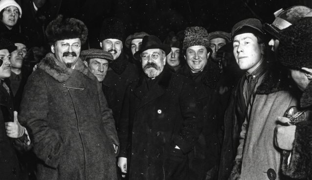 Fotografia histórica em preto e branco mostrando um grupo de homens em um ambiente noturno com neve caindo. Eles vestem casacos pesados e chapéus de inverno. Em destaque à esquerda está Leon Trotsky, sorrindo, com bigode, cavanhaque e um casaco com gola de pele. Ao centro está Lev Kamenev, usando óculos redondos, barba e um sobretudo escuro. A imagem possui textura granulada de filme antigo.