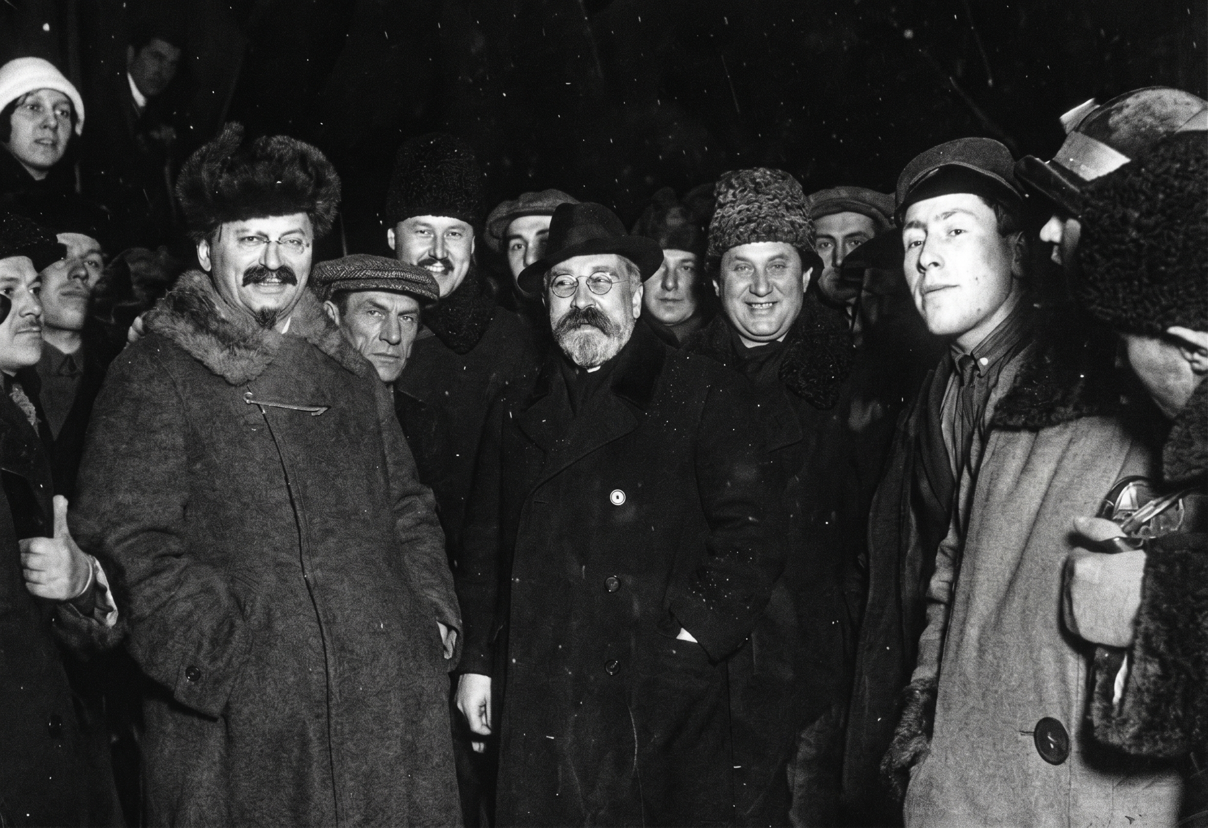 Fotografia histórica em preto e branco mostrando um grupo de homens em um ambiente noturno com neve caindo. Eles vestem casacos pesados e chapéus de inverno. Em destaque à esquerda está Leon Trotsky, sorrindo, com bigode, cavanhaque e um casaco com gola de pele. Ao centro está Lev Kamenev, usando óculos redondos, barba e um sobretudo escuro. A imagem possui textura granulada de filme antigo.
